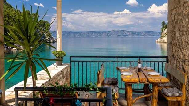 Una terraza con mesa de madera lista para comer, con vista al mar tranquilo y montañas distantes.