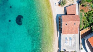 Vista aérea de una propiedad costera con playa, un edificio de techo rojo y aguas turquesas cristalinas.
