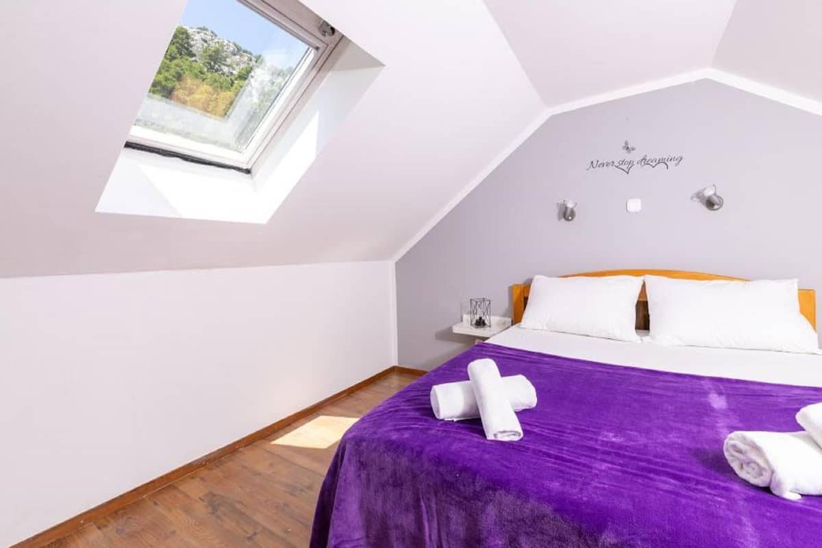 Un dormitorio con tragaluz, cama con ropa de cama blanca y naranja, una manta morada y una toalla.