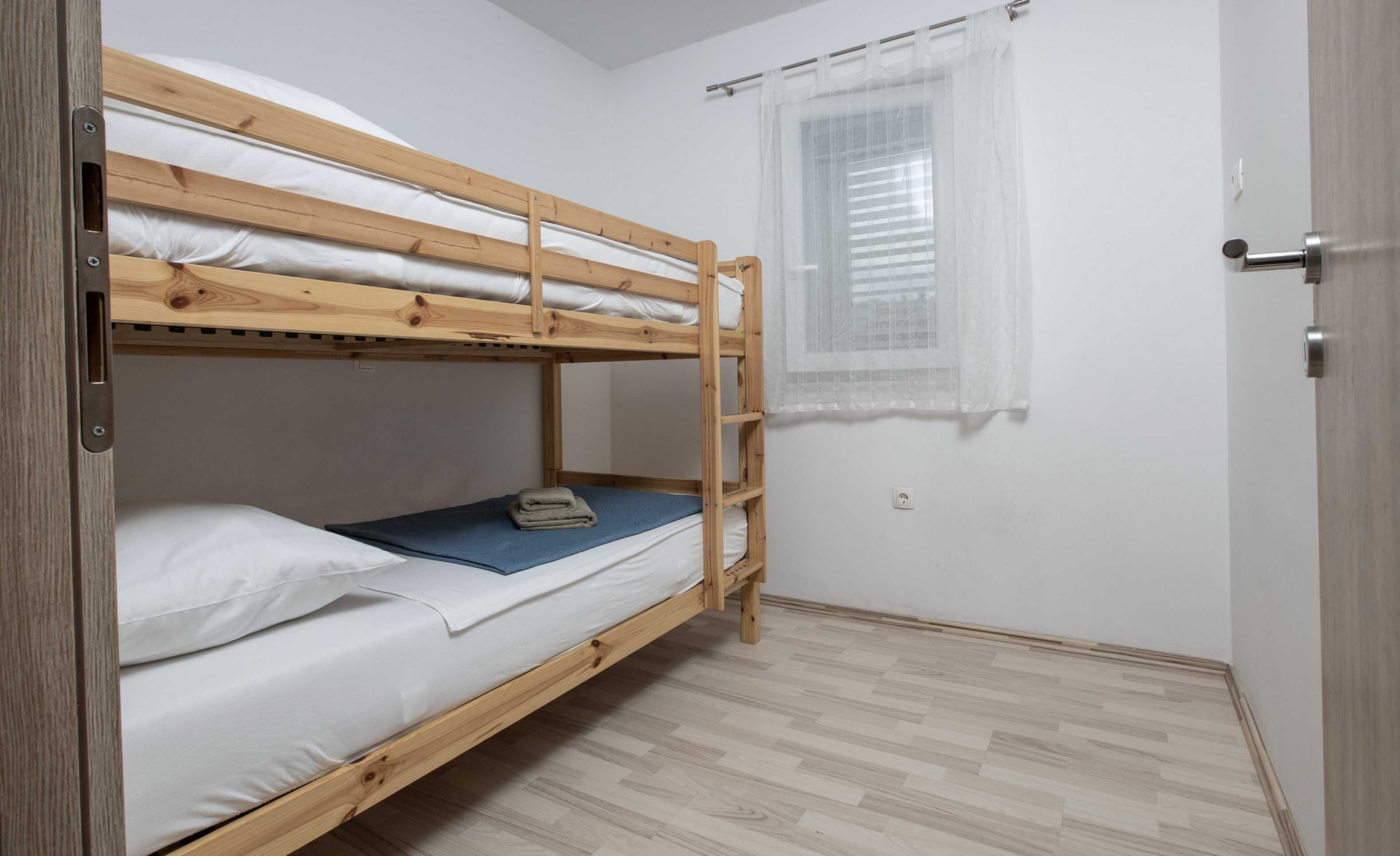 A single bunk bed room with a wooden frame, white bedding, a small window with sheer curtains, and a door with a handle.