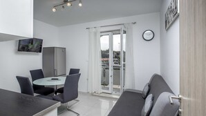 Een moderne woonkamer met een bank, een eettafel met stoelen, een flatscreen-tv aan de muur, een koelkast en een schuifdeur naar een balkon.