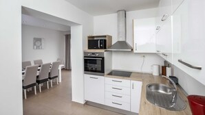 Een moderne keuken met witte kastjes, een magnetron, oven en een spoelbak. Er is een eethoek met een tafel en stoelen zichtbaar op de achtergrond.