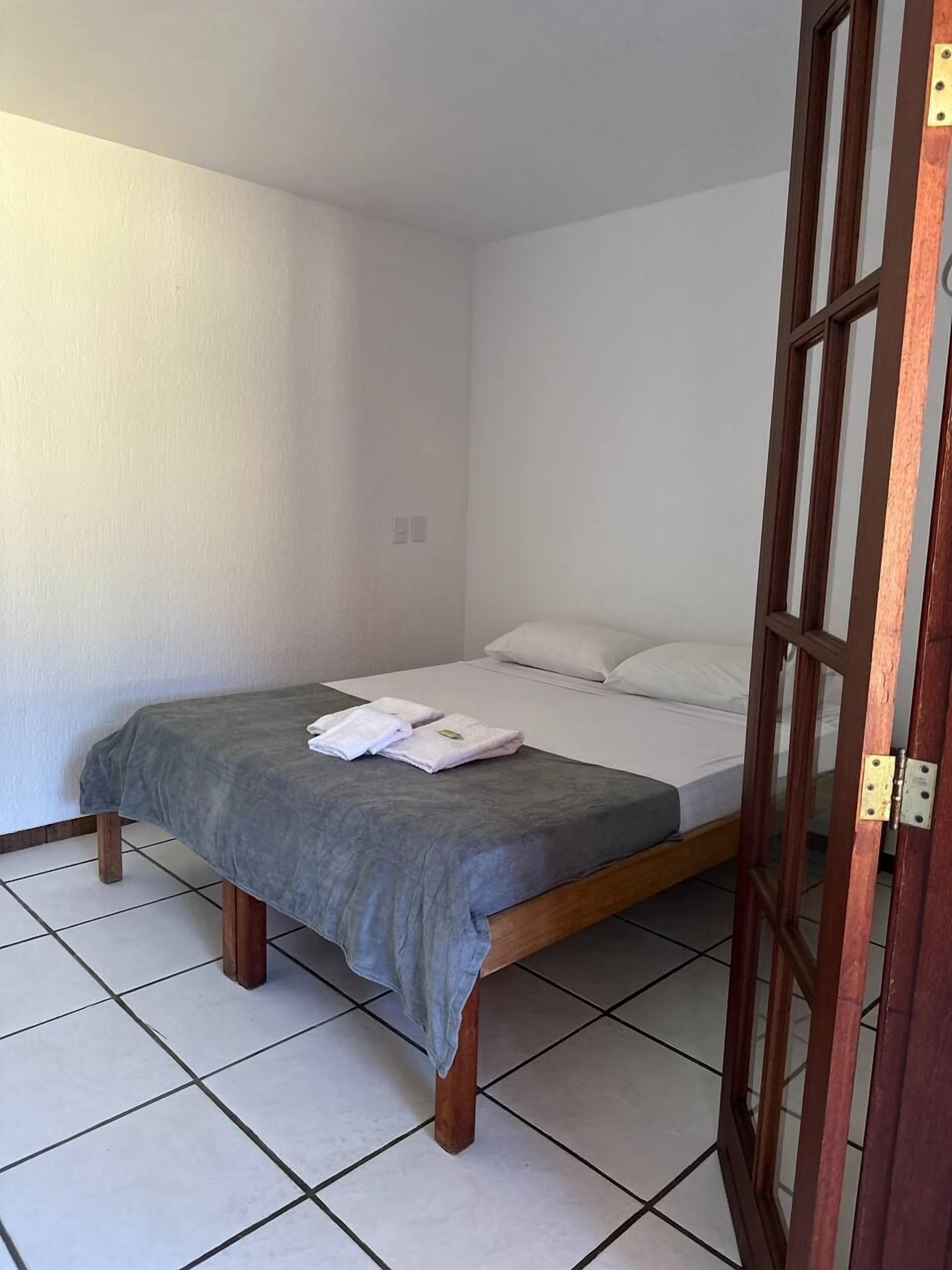 Quarto individual com cama de madeira, roupa de cama branca e manta cinza.