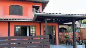 A building with a red exterior, a wooden bench, and a covered patio area with chairs and tables. - Pousada Encanto Torrely (Búzios)