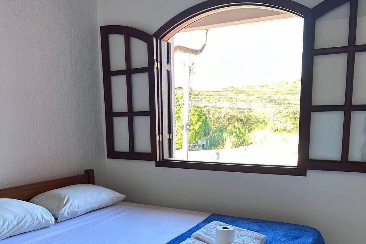 Un lit simple avec une couvre-lit bleue et des oreillers blancs, une fenêtre dont les volets sont ouverts, et une vue sur la verdure à l’extérieur.