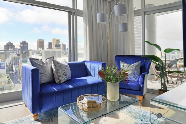 Un salon moderne avec un canapé bleu, une table basse en verre et une vue sur le paysage urbain.