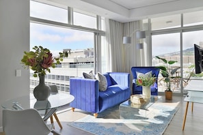 Un salon moderne avec un canapé bleu, une table basse en verre et une vue sur la ville grâce à de grandes fenêtres.