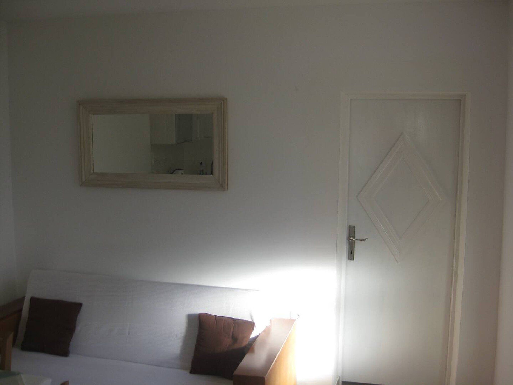 Una habitación con una cama blanca, una almohada marrón, un espejo en la pared y una puerta con un diseño en forma de rombo.