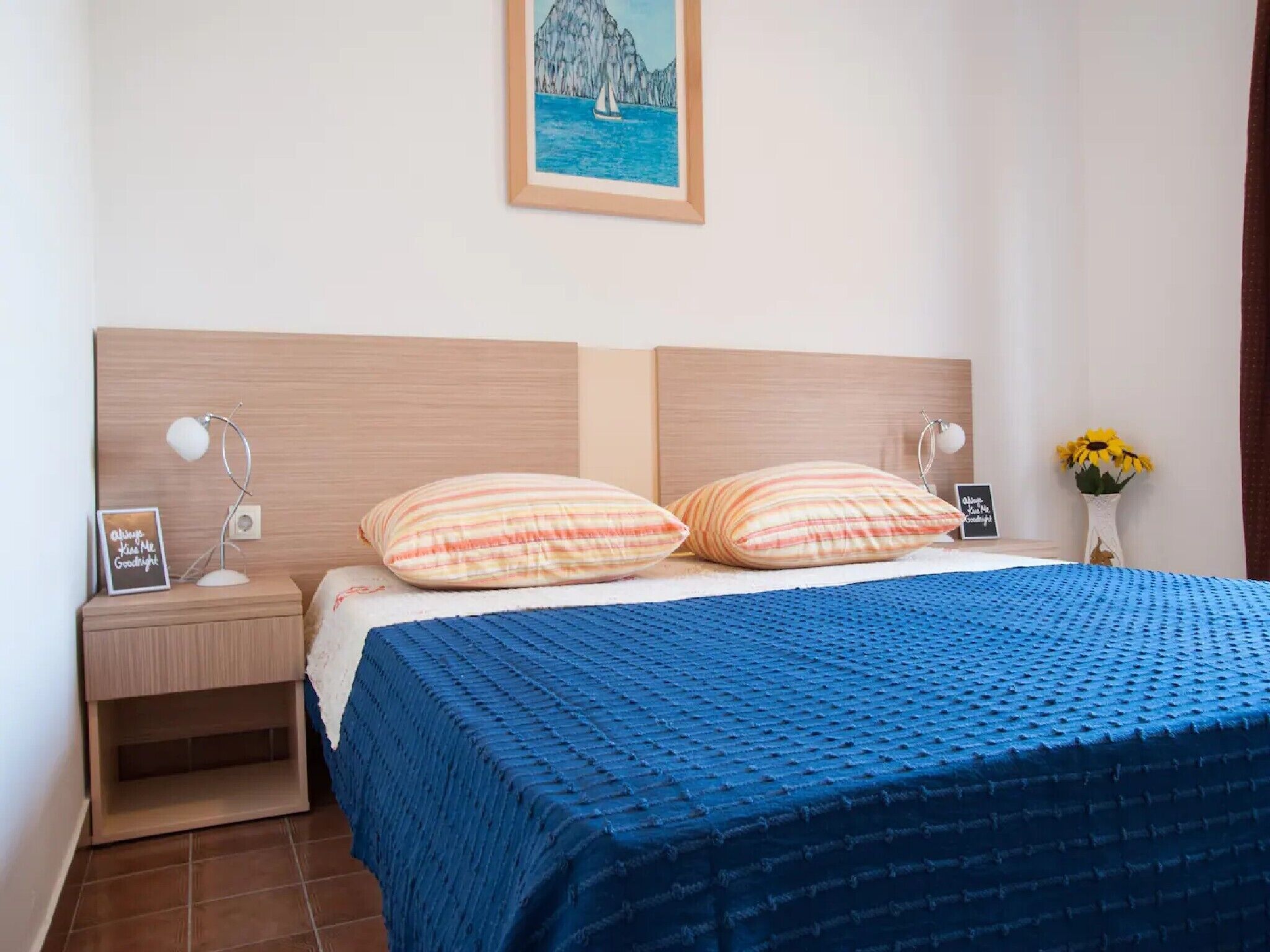 A neatly made bed with a blue textured bedspread, two striped pillows, and a wooden headboard. A small bedside table with a lamp and a framed picture is visible.