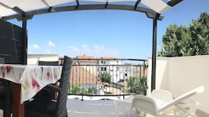 Ein überdachter Außenbereich mit Tisch und Stühlen, im Hintergrund Gebäude und Bäume.