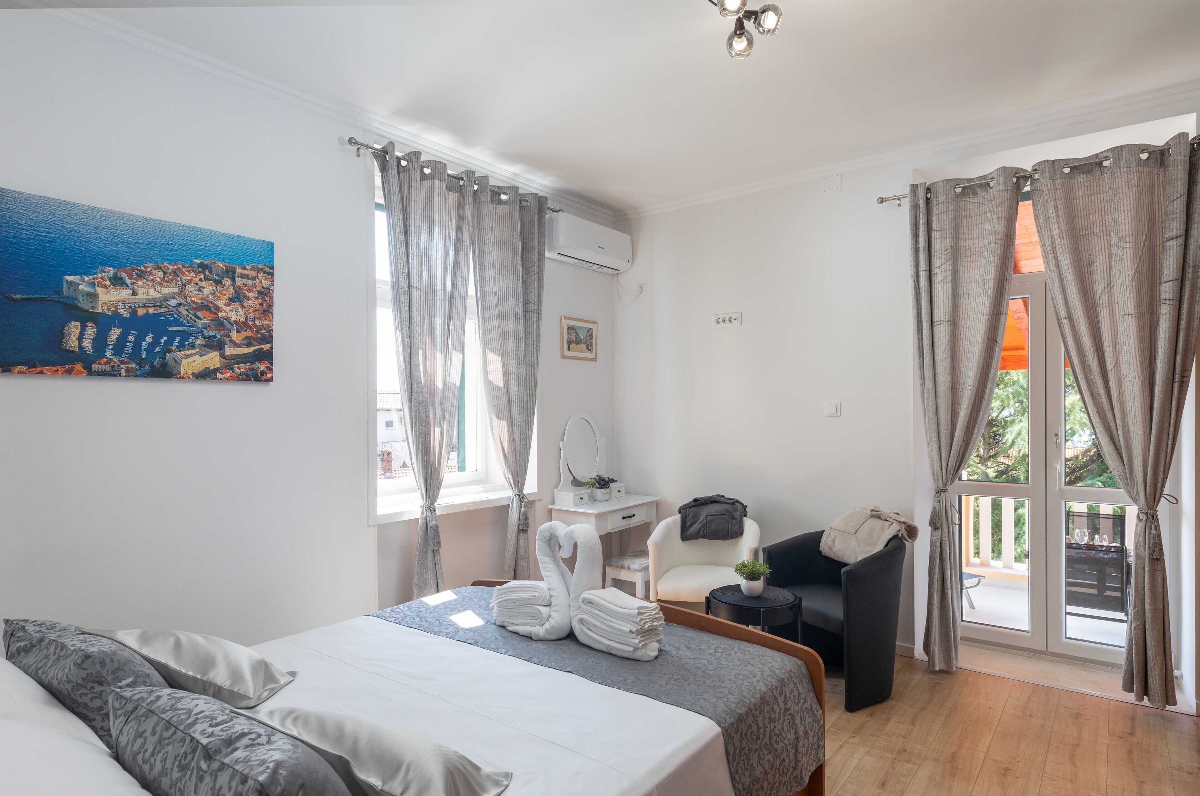 A hotel room with a bed, a desk, a chair, a balcony, and a painting of a coastal town.