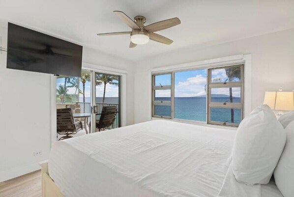 A bedroom with a bed, a ceiling fan, a TV mounted on the wall, and a view of the ocean through large windows.