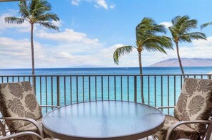A balcony with a glass table and chairs, overlooking a clear blue sea with palm trees and a distant mountain.