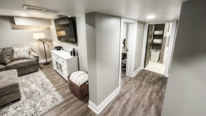 A modern living room with a sofa, a small table, a lamp, and a TV. There is a hallway leading to a laundry area.