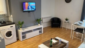 A modern living room with a flat-screen TV, a washing machine, a dining area with a table and chairs, and a small plant on a side table. - Cozy and Central City Apartment in Liverpool (Liverpool)