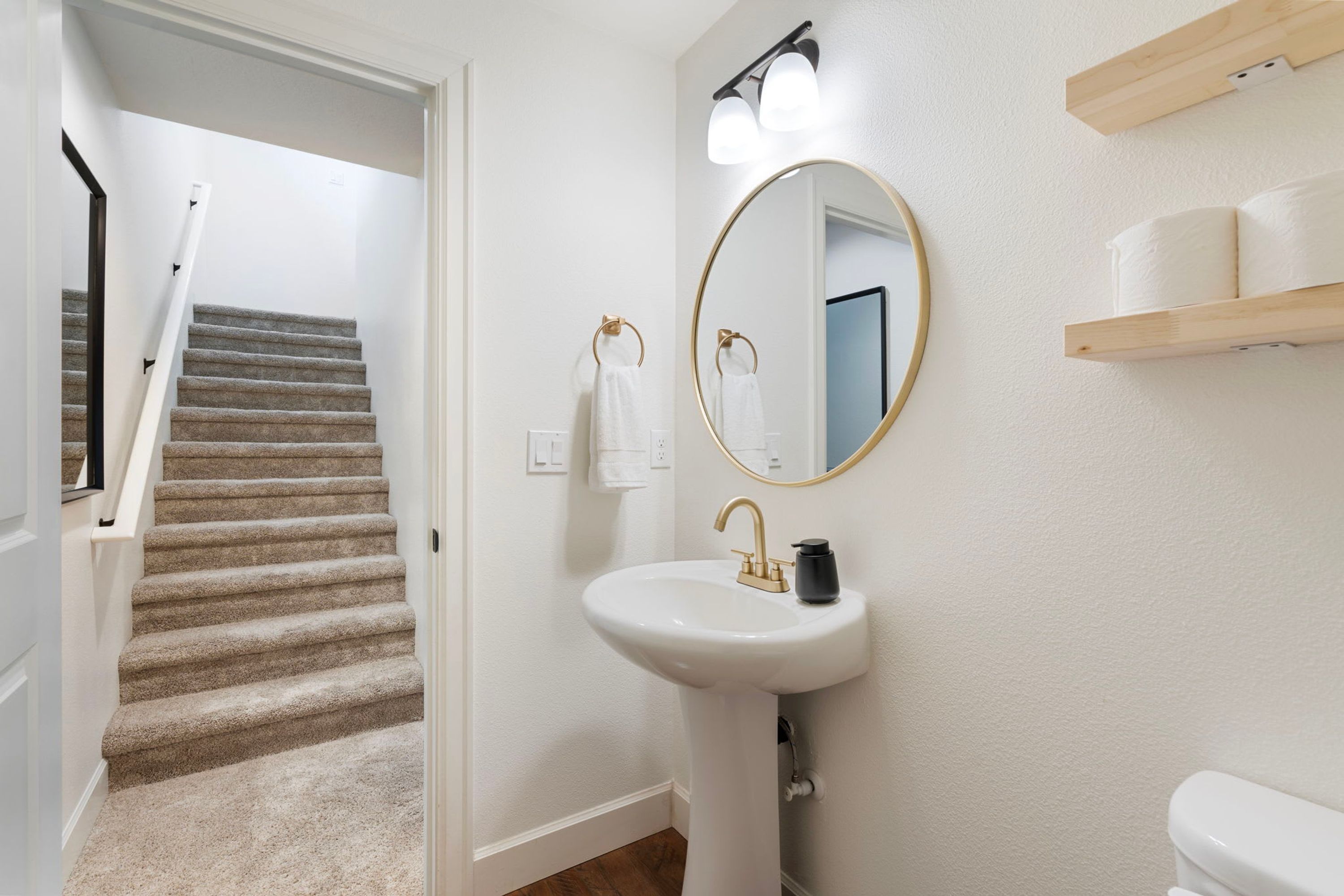 A bathroom with a white sink, a round mirror, and a toilet. There is a wooden shelf above the toilet and a staircase leading to another room.