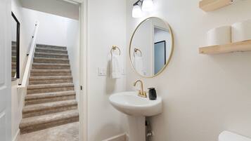 A bathroom with a white sink, a round mirror, and a toilet. There is a wooden shelf above the toilet and a staircase leading to another room.