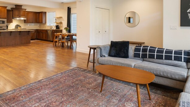 Amplia sala de estar con sofá, mesa de centro y alfombra estampada. Se ve una cocina con armarios de madera y un comedor con sillas al fondo.