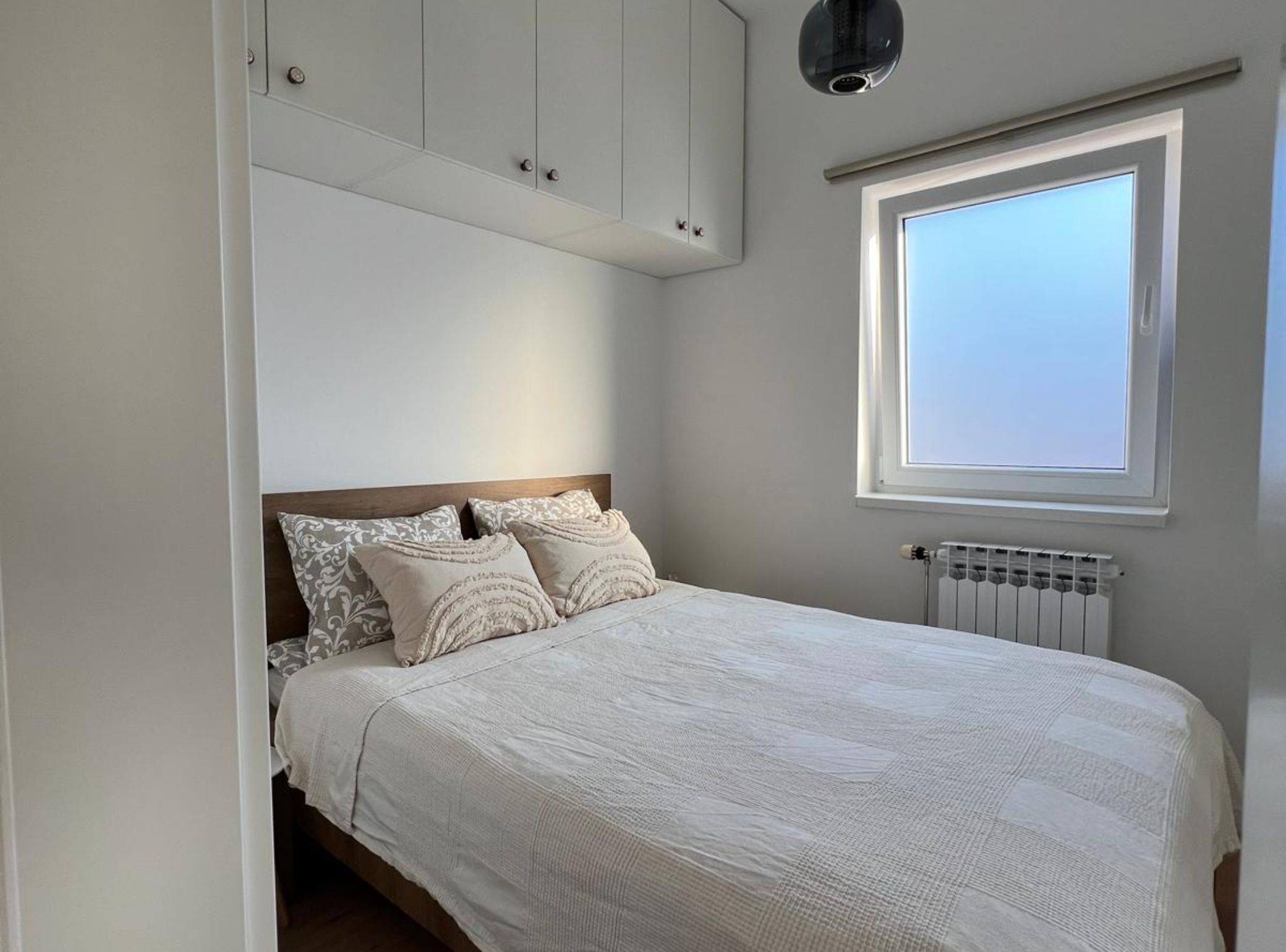 Ein Schlafzimmer mit Bett, Fenster, Heizkörper und einer Deckenleuchte.