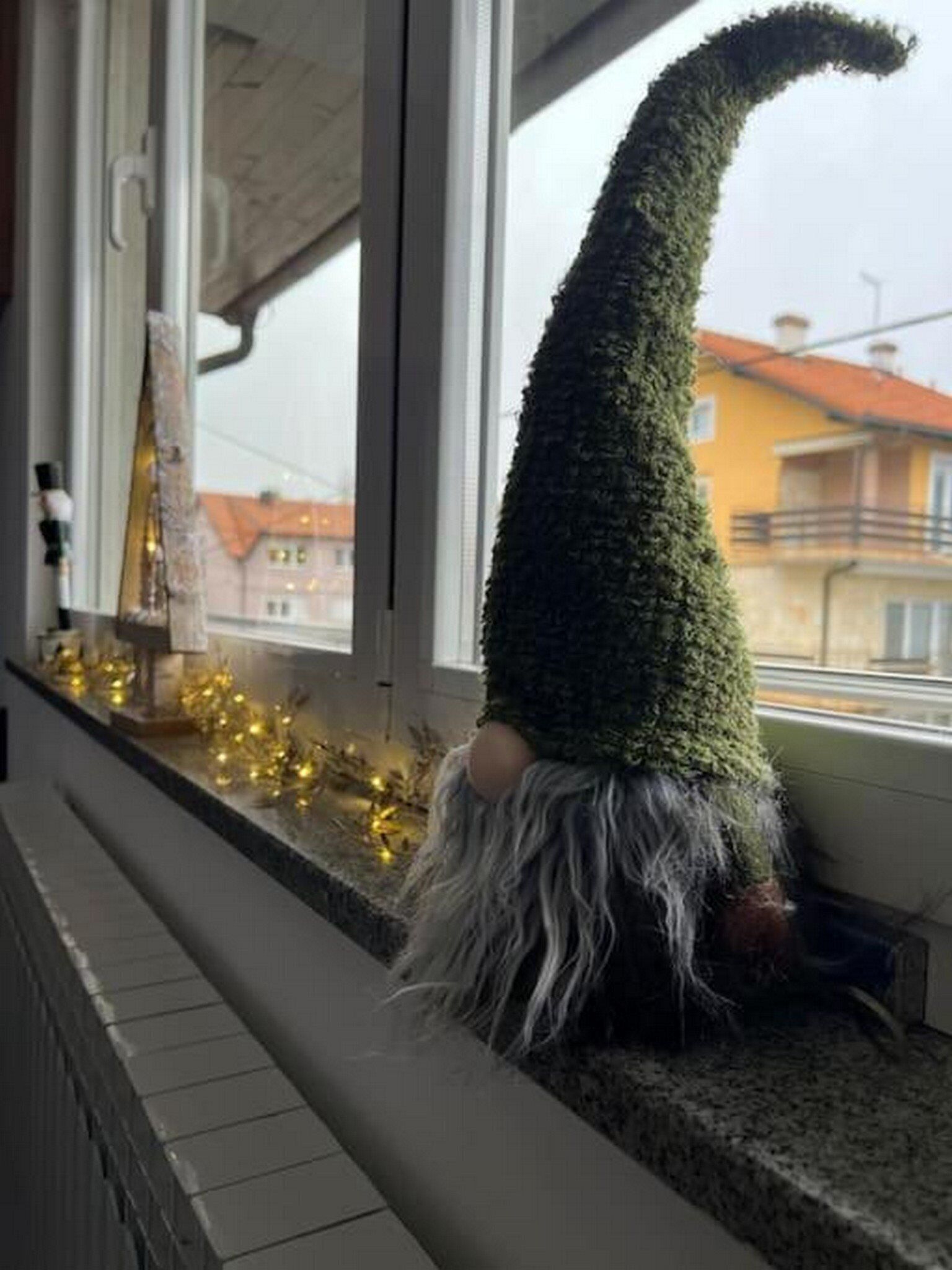 A gnome figurine with a pointed hat and a long beard, placed on a windowsill with a view of buildings outside.