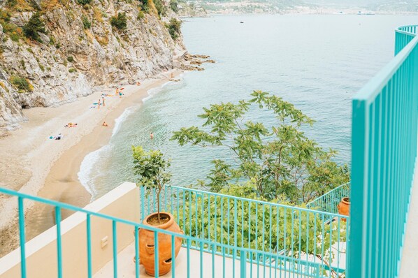 A coastal view from a balcony with a turquoise railing, overlooking a sandy beach with people and a calm sea.