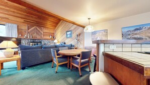 A cozy living area with a wooden ceiling, a stone fireplace, a dining table with chairs, and a kitchen area with wooden cabinets. - Large Rustic Home. Private Washer/Dryer, Centrally Located. Pool On-site (Mammoth Lakes)