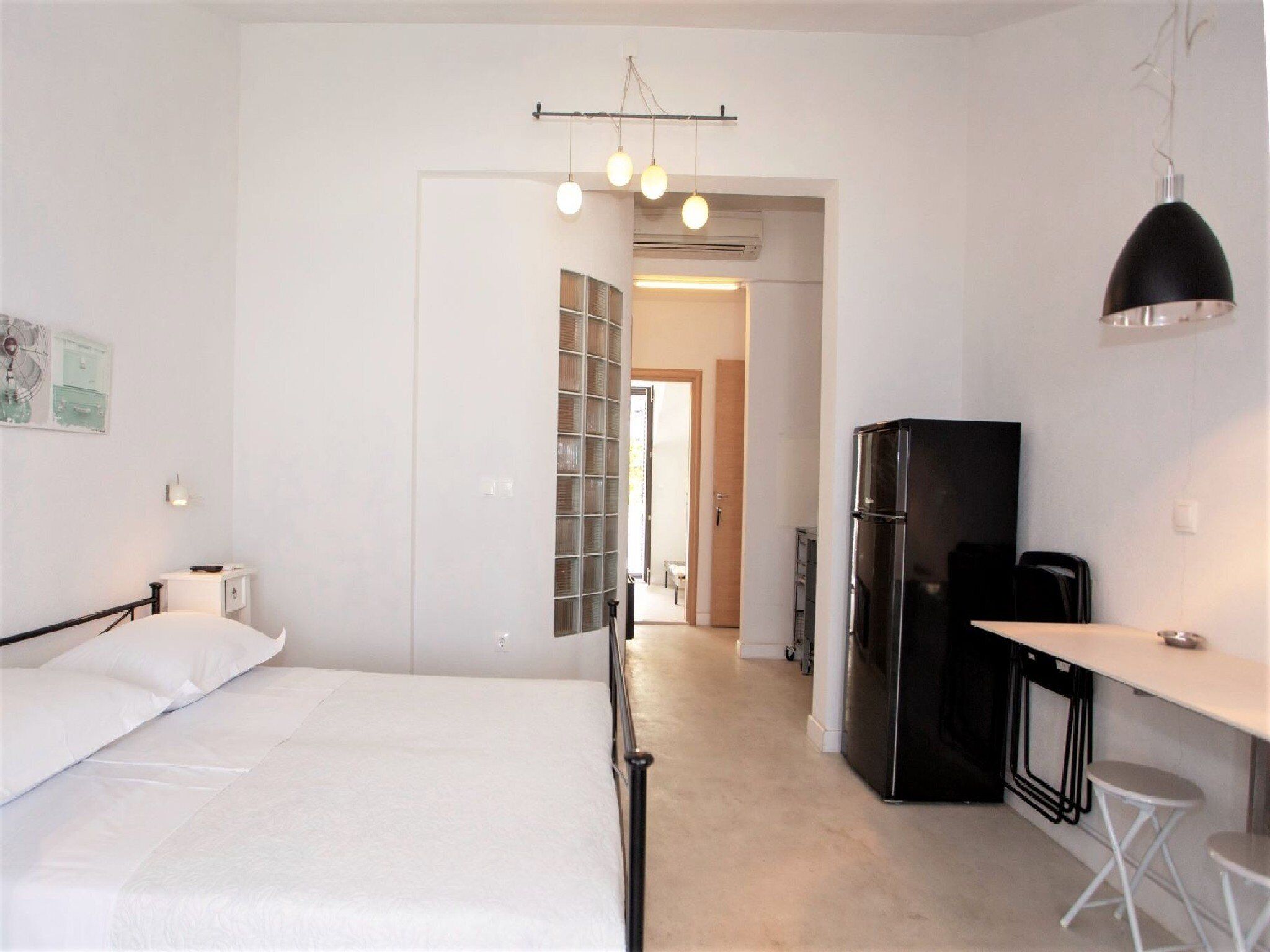 A compact hotel room with a bed, a small kitchenette, a dining area with stools, and a hallway leading to another room.