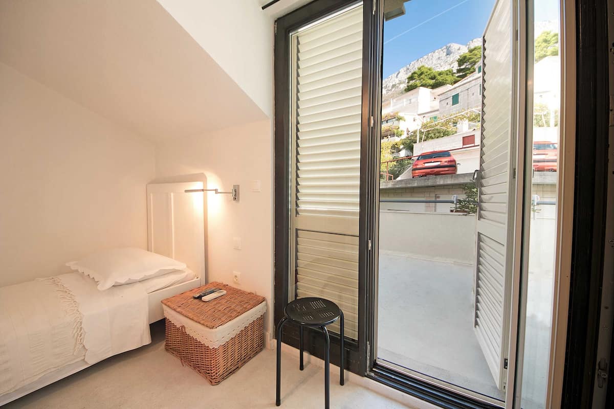 A small bedroom with a bed, a nightstand, a lamp, a wicker basket, and a sliding glass door leading to an outdoor area.