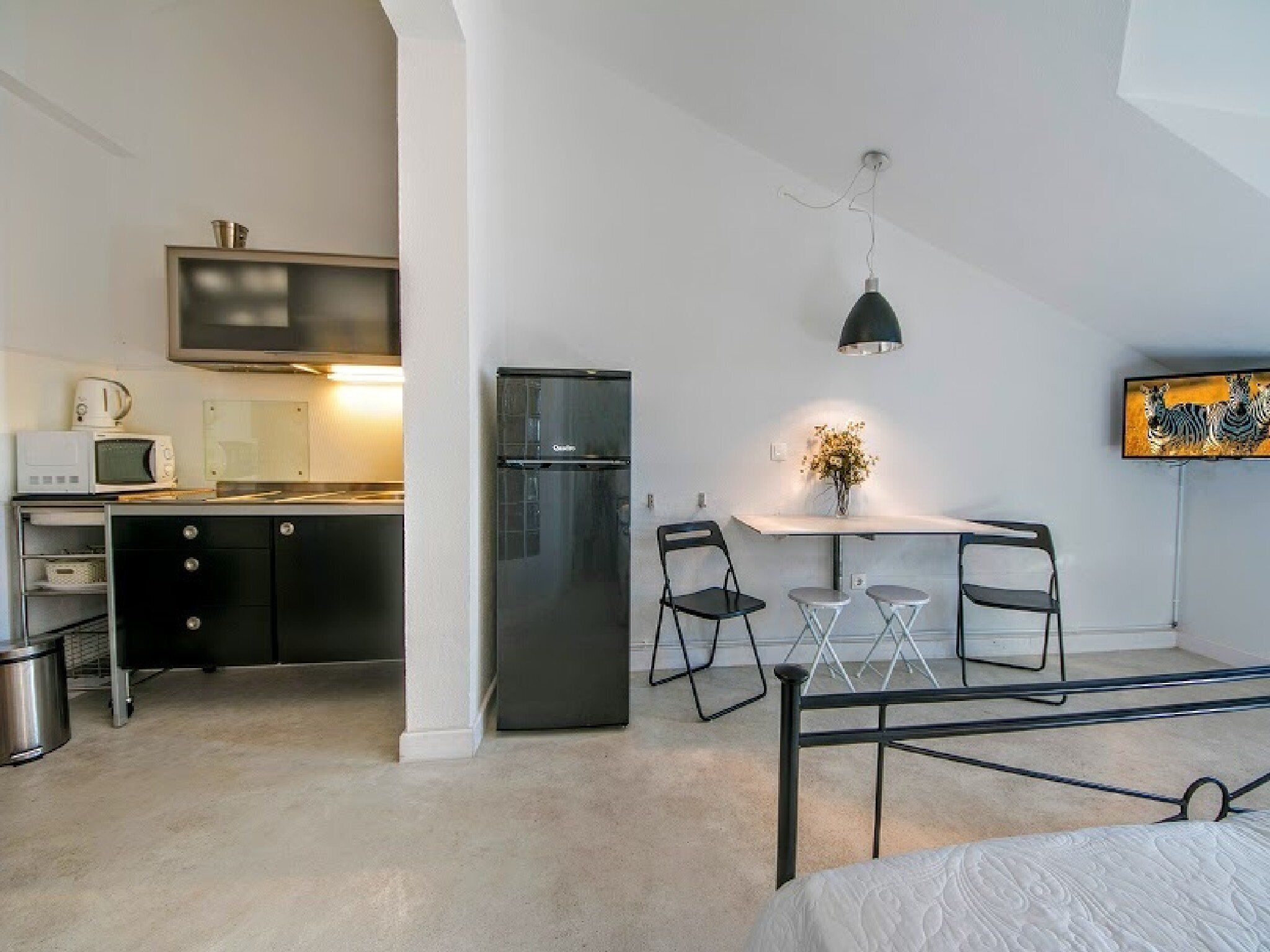 A compact bedroom with a bed, a small kitchenette, a dining table with chairs, and a TV mounted on the wall.