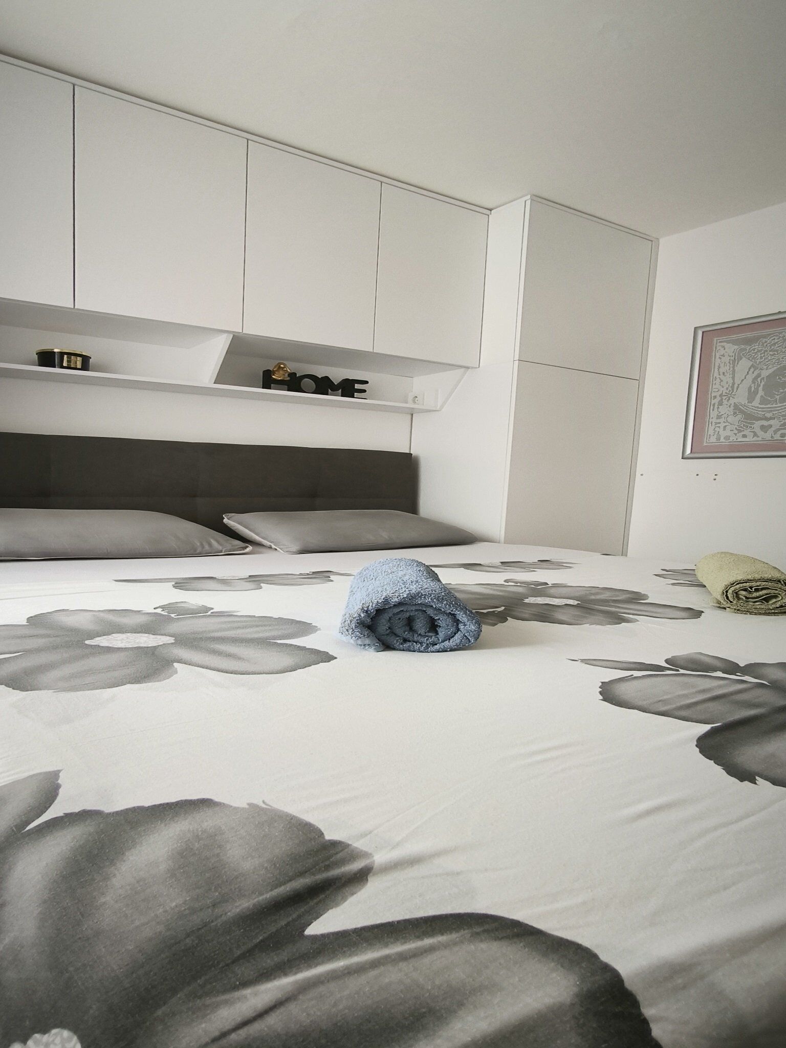 A bedroom with a bed, a headboard, a shelf with a 'HOME' sign, and a framed picture on the wall.