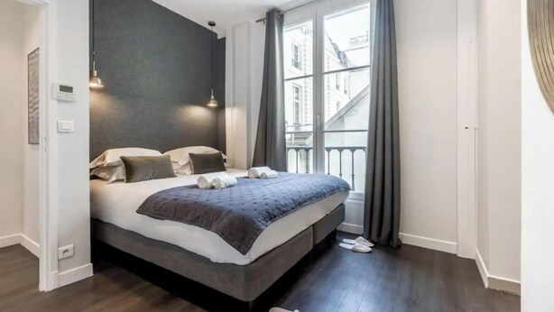 A modern bedroom with a large bed, dark walls, and a view of buildings through a window. - ELEGAN PARISIAN APARTMENT-LUXURY,COMFORT & SPECTACULAR VIEWS AWAIT– RUE MONSIGNY (Paris)