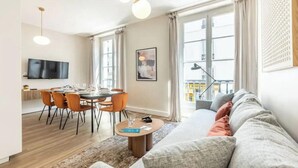 A modern living room with a sofa, dining table, and chairs. There is a TV mounted on the wall and a window with curtains. - ELEGAN PARISIAN APARTMENT-LUXURY,COMFORT & SPECTACULAR VIEWS AWAIT– RUE MONSIGNY (Paris)