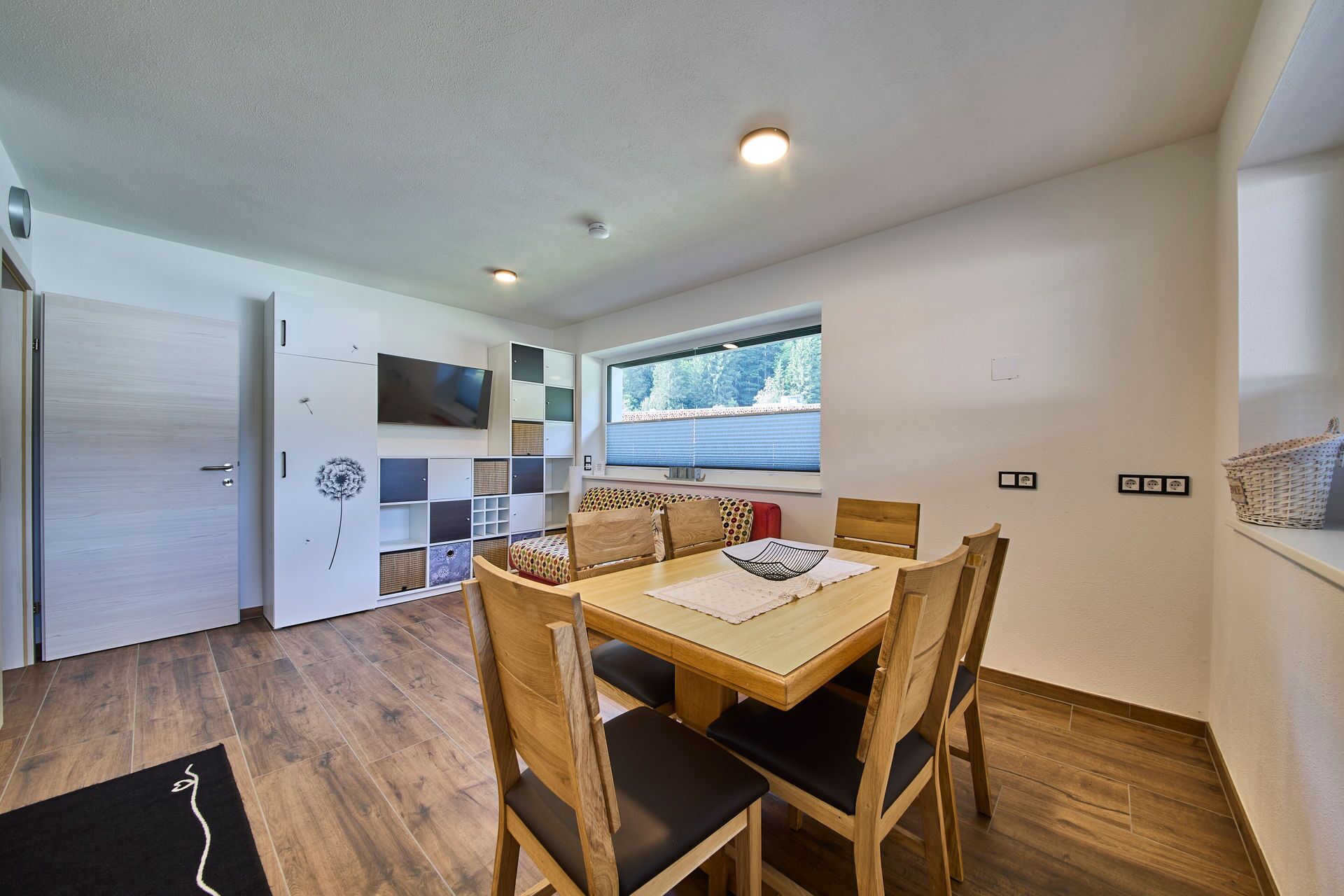 A dining area with a wooden table and chairs, a television mounted on the wall, and a window with blinds.