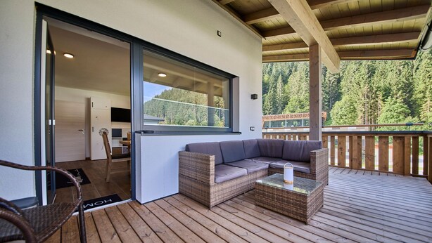 A covered outdoor area with a wicker sofa and a glass table, a wooden deck, and a view of a forested area. - Haus Joan - Apartments (Saalbach-Hinterglemm)