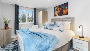 Ein modernes Schlafzimmer mit einem großen Bett, einem Nachttisch mit Lampe, einem Fenster mit Vorhängen und einem Bild an der Wand.