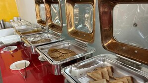 A buffet setup with various food items in metal trays, including sandwiches and cookies, on a red table with disposable plates and utensils. - Opulent Inn By Lime Tree Hotels (Greater Noida)