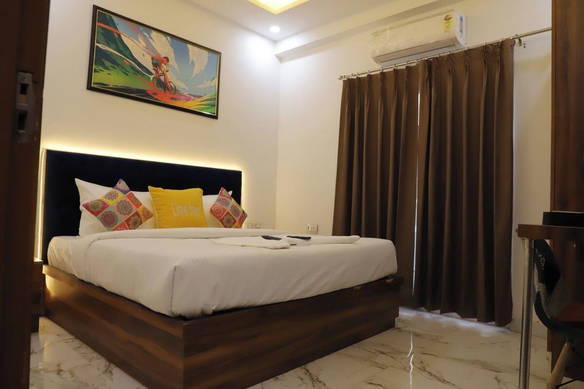 A hotel room with a bed, a headboard, a painting, a curtain, and an air conditioning unit.