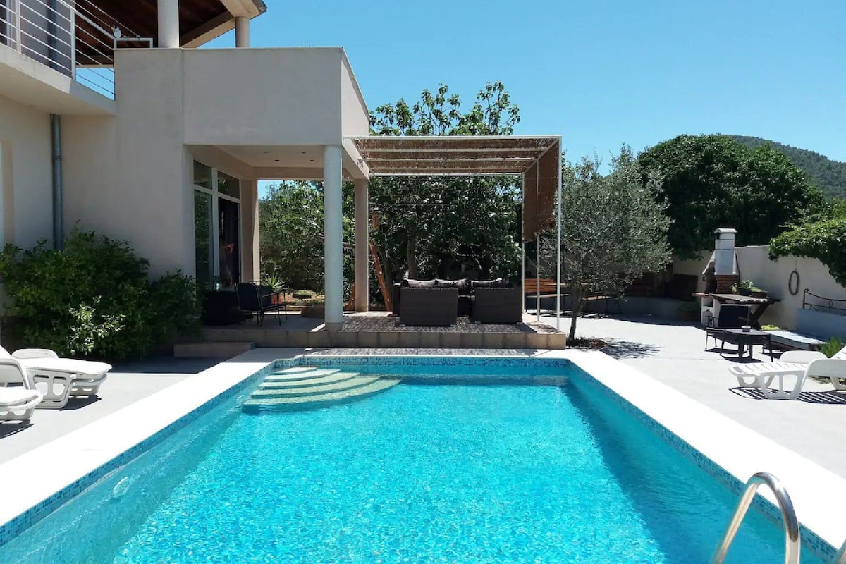 Un área de piscina moderna al aire libre con un pérgola, asientos y una zona de barbacoa.