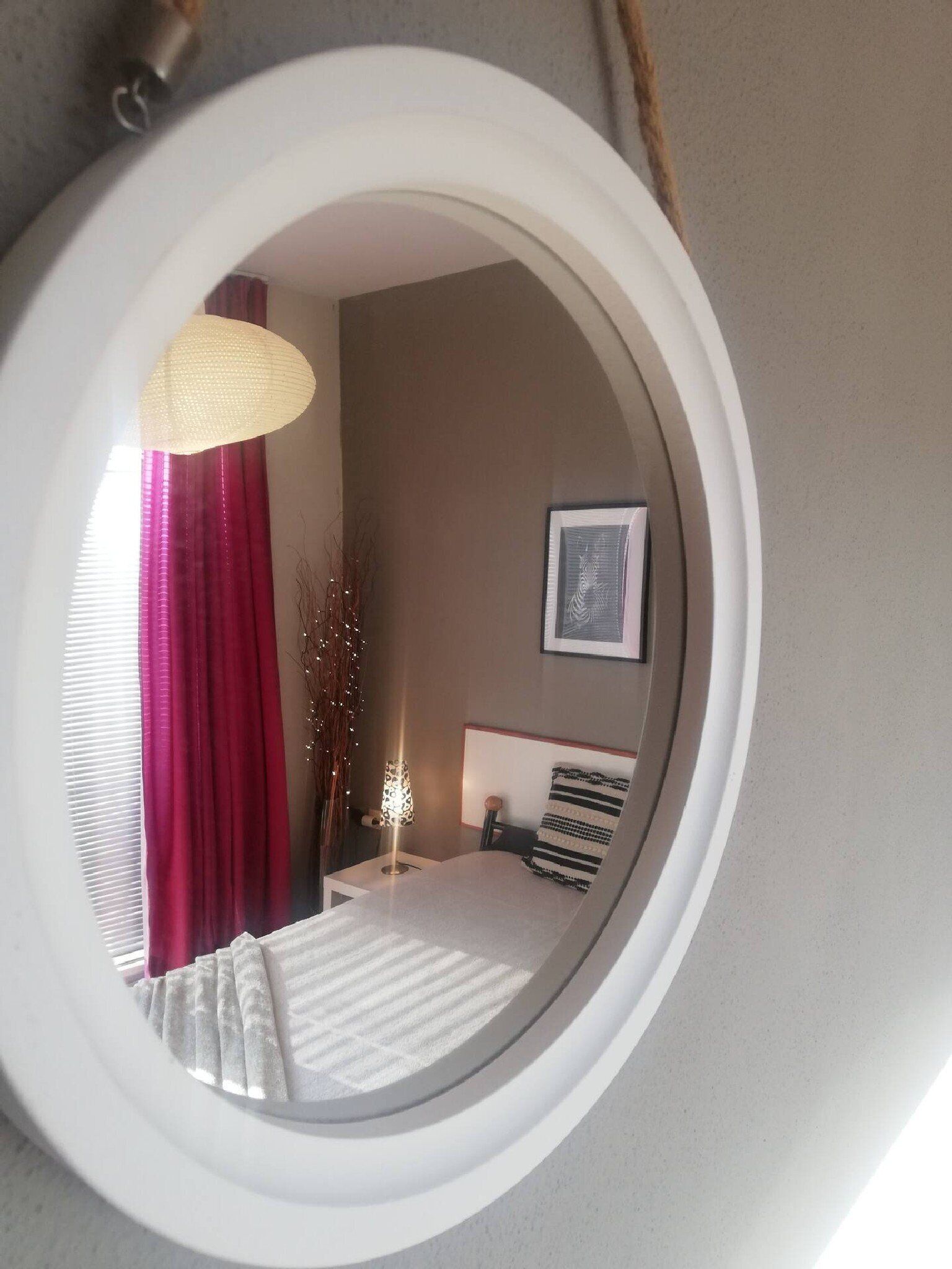 A bedroom with a round mirror, a bed with striped bedding, a bedside table with a lamp, and a framed picture on the wall.