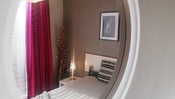 A bedroom with a round mirror, a bed with striped bedding, a bedside table with a lamp, and a framed picture on the wall.