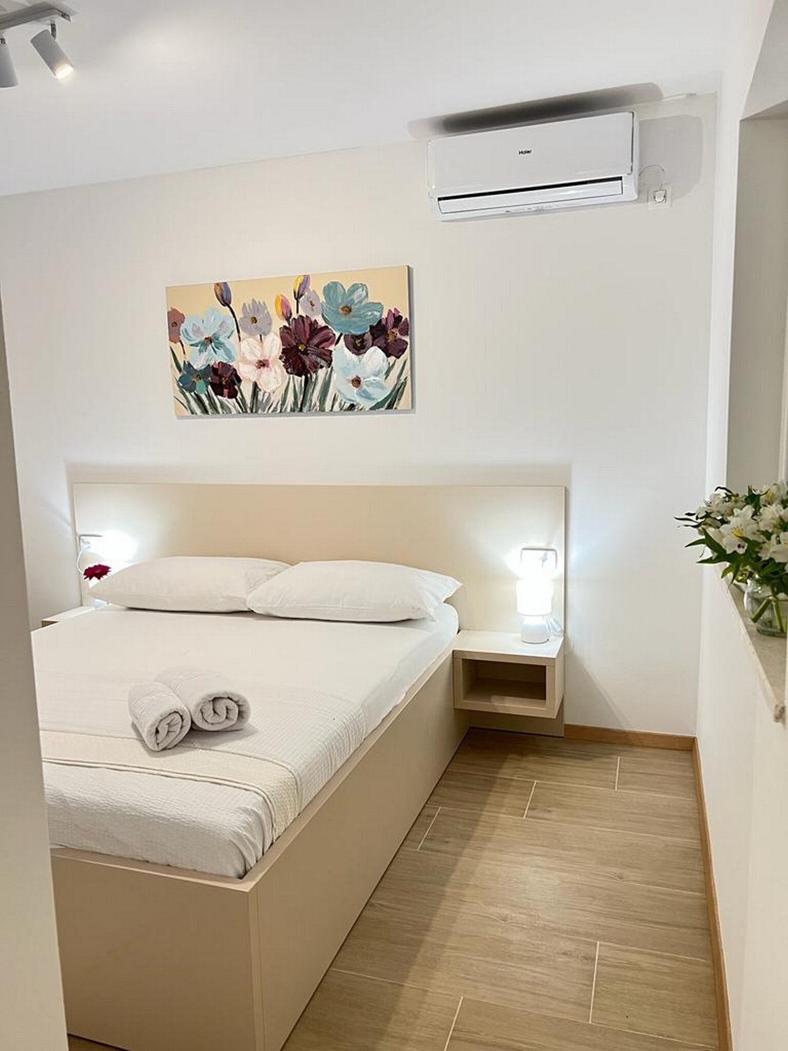 A hotel room with a bed, a nightstand, an air conditioning unit, and a floral painting on the wall.