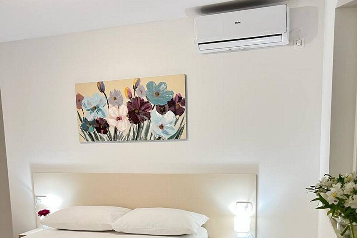A hotel room with a bed, a nightstand, an air conditioning unit, and a floral painting on the wall.