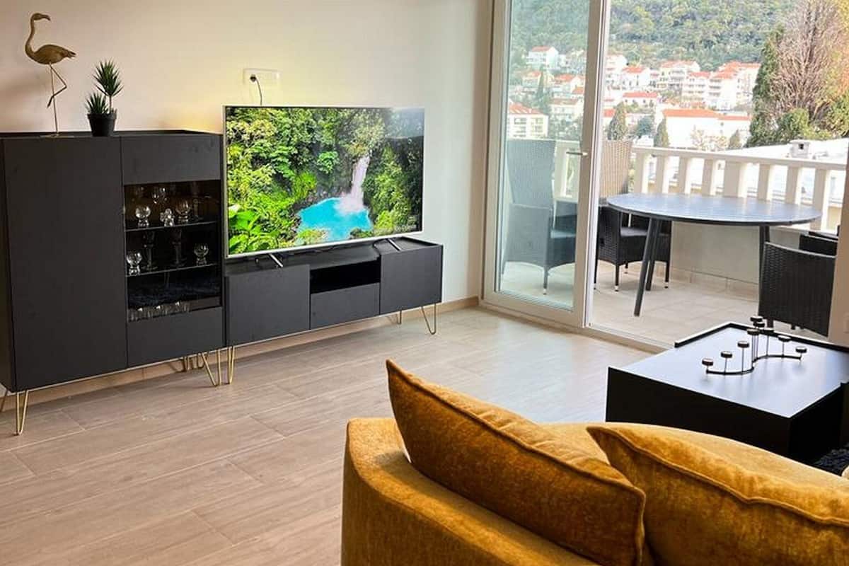 A modern living room with a flat-screen TV, a sofa, a dining area, and a balcony with a view of buildings.