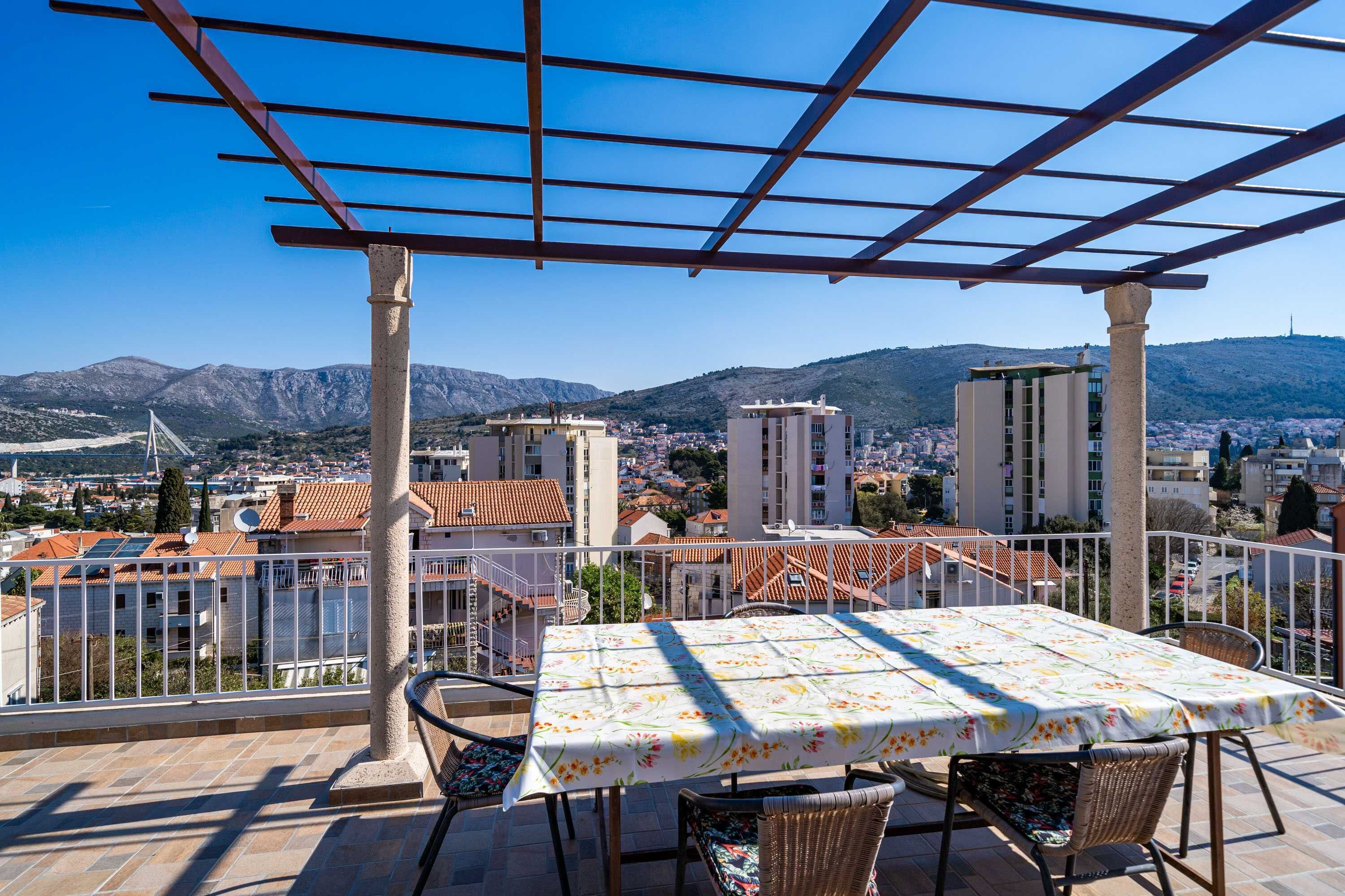 A terrace with a table and chairs, overlooking a cityscape with buildings and mountains.