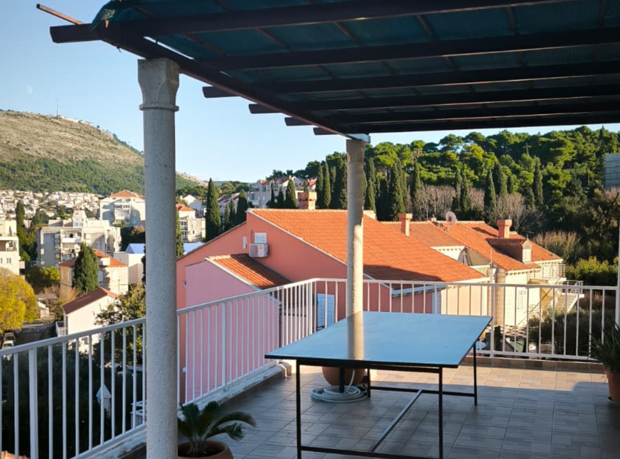 A terrace with a table and chairs, overlooking a residential area with red-roofed houses and a hill in the background.