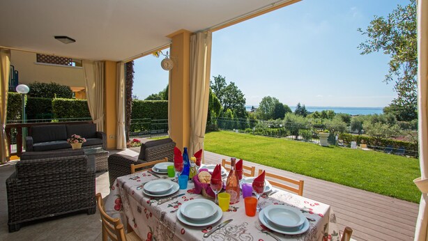 An outdoor dining area with a table set for four, surrounded by wicker furniture, and a view of a well-maintained garden and sea. - Crocus 1 con piscina by Wonderful Italy (Soiano del Lago)