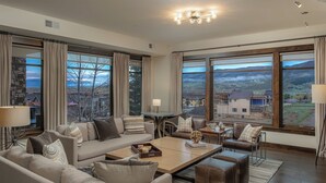 A spacious living room with a large sofa, coffee table, and dining table. There are large windows with curtains, providing views of a mountainous landscape.
