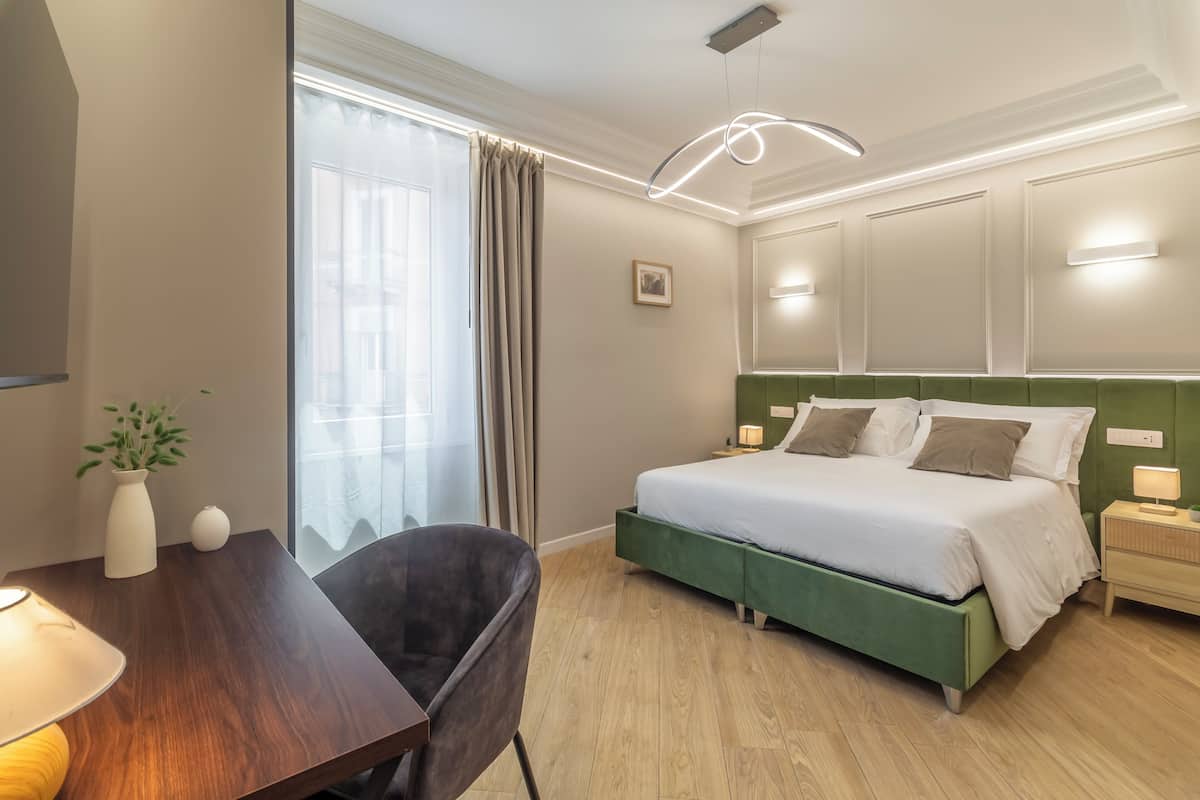 Una habitación de hotel con cama, mesita de noche, lámpara, silla y ventana con cortinas.