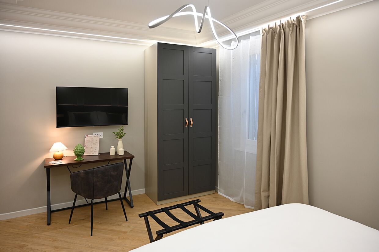 A bedroom with a bed, a desk with a lamp, a TV mounted on the wall, a wardrobe, and a window with curtains.