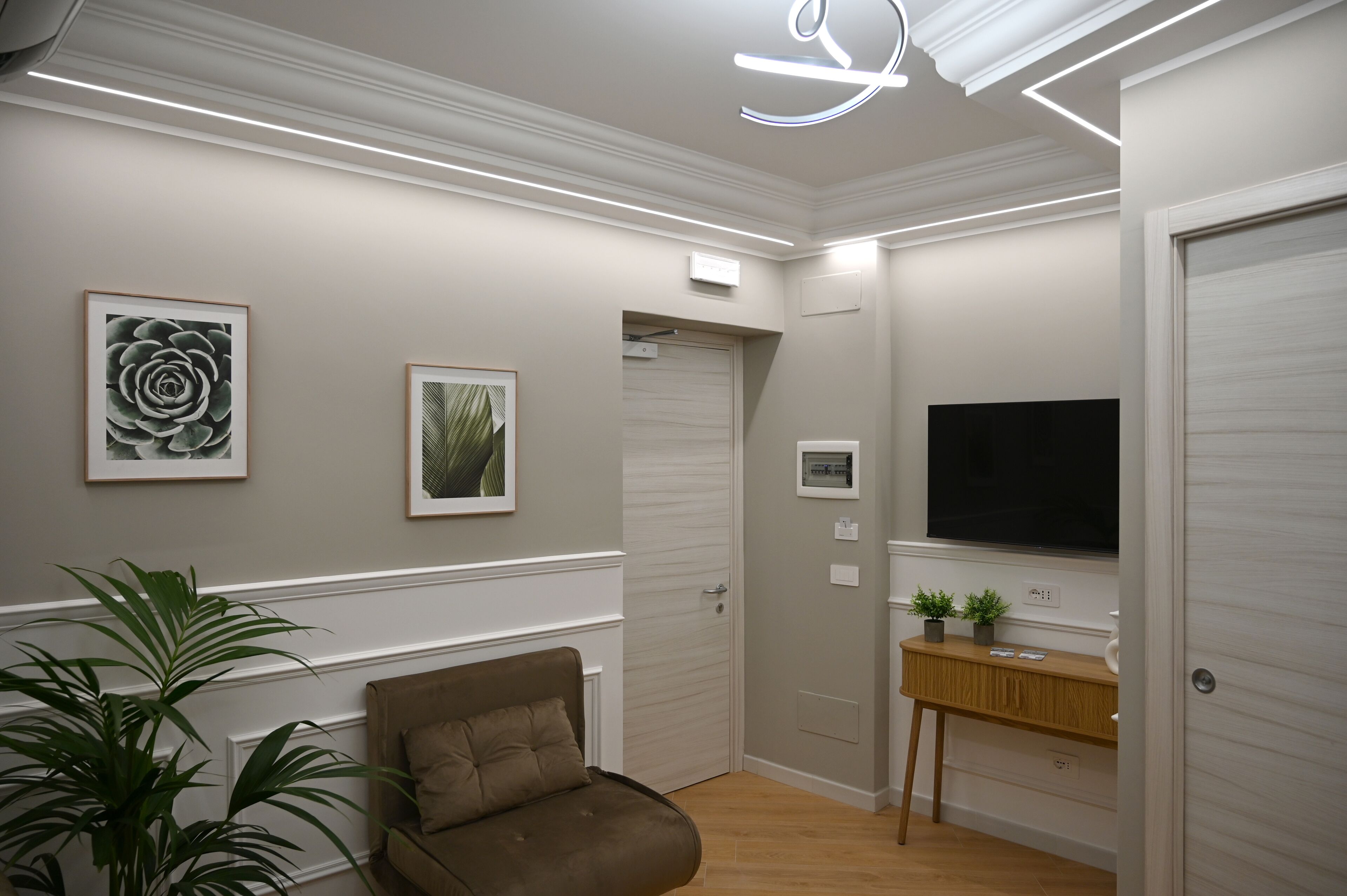 A modern hallway with a brown sofa, framed floral artwork, a wooden console table, and a large TV on the wall.